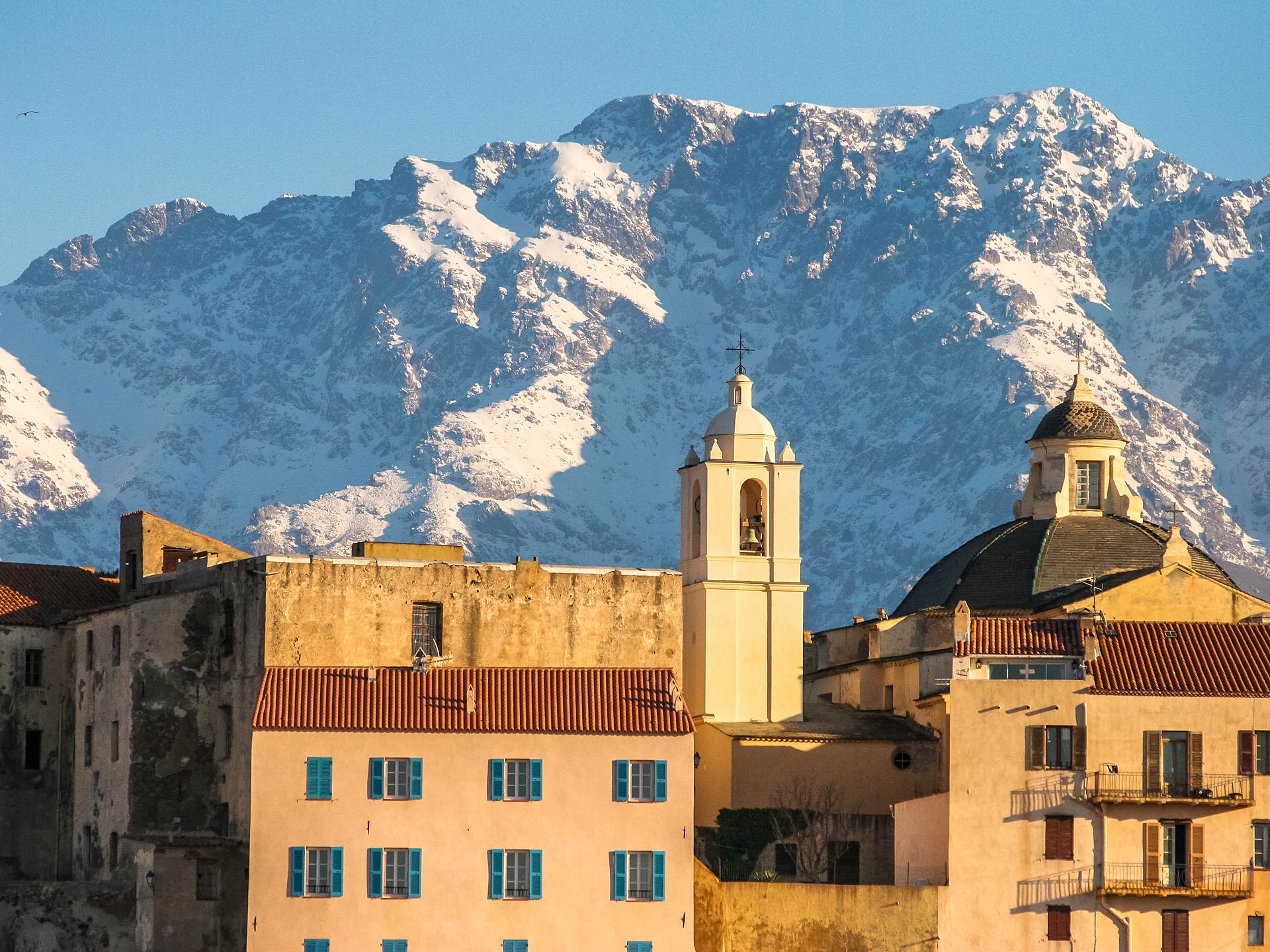Village corse aux maisons pastel et clocher blanc, avec montagnes enneigées en arrière‑plan